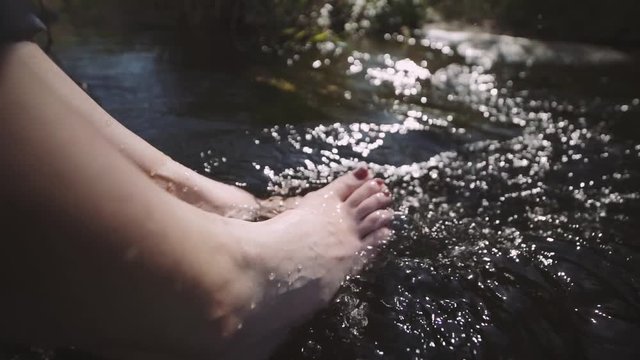 Woman relaxes by river sitting on edge of rock, swing one's feet on water flow. Girl having fun during vacation on the beach at morning. Slow motion 