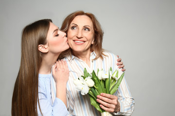 Young woman greeting her mother with bouquet on light background