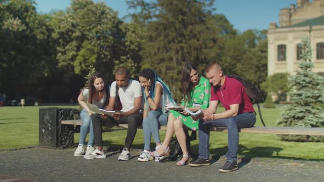 Group of diverse college students talking after classes sitting on bench. Multi ethnic classmates intending to spend freetime at campus together while upset female friend must go home after phone call