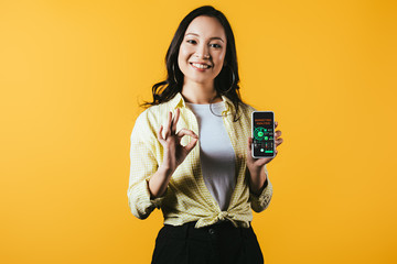 smiling asian girl showing ok sign and smartphone with marketing analysis, isolated on yellow