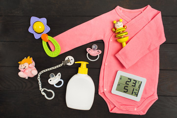 Baby accessories and digital thermo-hygrograph on dark wooden background. Pink bodysuit, two rattles, two pacifiers, toy and soap with hygrometer and thermometer for baby health