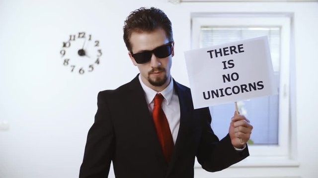 Businessman holds a sign There Is No Unicorns 4K. Medium shot of a male person in focus dressed up nicely with a red tie. Wall with clock and window in the background.