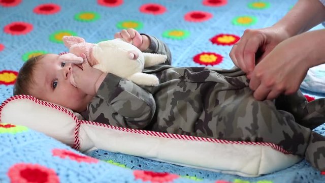 Mom removes clothes from a baby. Baby is playing with a soft toy while lying on a bed. Clothes are being removed from him.