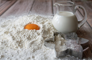 Flour, milk in with a glass jug, orange chicken yolk and a baking dish star on brown wooden table boards closeup