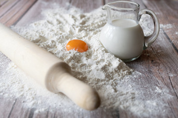 Scattered flour, rolling pin, milk in a glass jug, orange chicken yolk and a baking dish star on brown wooden table boards