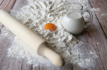 Flour, rolling pin, milk in with a glass jug and orange chicken yolk on brown wooden table boards close-up