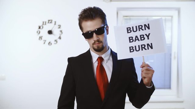 Businessman hold sign Burn Baby Burn 4K. Medium shot of a male person in focus dressed up nicely with a red tie. Wall with clock and window in the background.