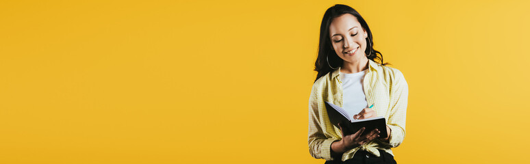 smiling asian girl writing in notebook with pen, isolated on yellow