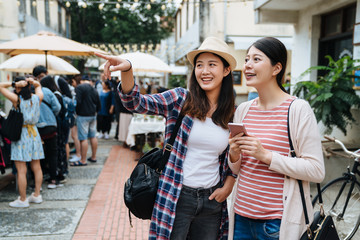 Fototapeta premium Young asian women tourists walking shopping in street food market in taipei taiwan. girls looking around at stand shops. female showing sister with cellphone point in outdoor holiday selling place.