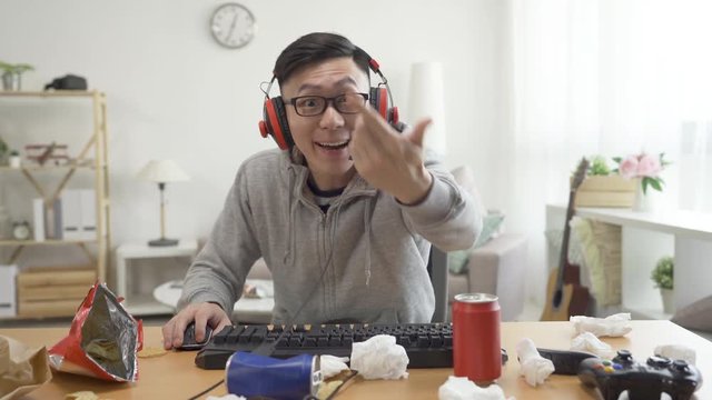 nervous asian man using desktop computer wear headset at home playing online video game. intense male talking on headphones with enemy on internet. teenage indoorsy at messy home on summer holidays.