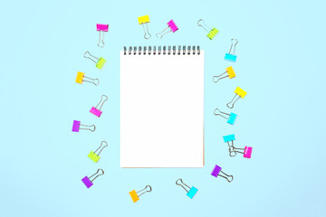 Back to school background with notepad, colorful paper clips on pastel blue backdrop. Flat lay, top view, copy space.