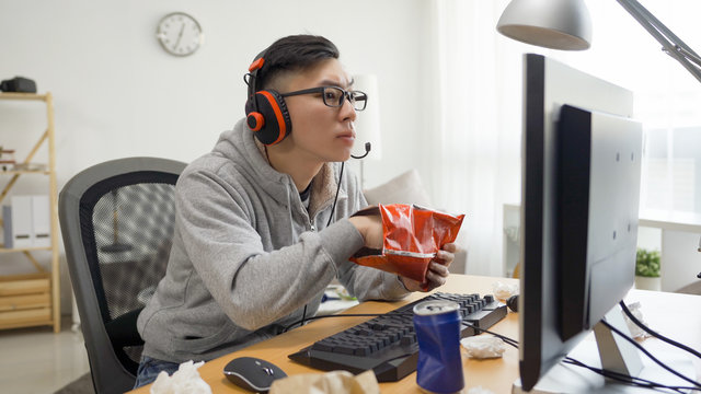 Asian Chinese Man Hand Eating Fast Food Bag Potato Chips With Soda Can While Having Fun Leisure Spend Time On Computer Game. Unhealthy Junk Food Lifestyle Concept. Otaku Watching Anime Summer Break