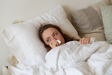 Sick young woman lying in bed covered with heavy blanket holding handkerchief