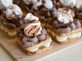 Portion cakes of meringue and cocoa butter cream in square form on a wooden stand, a piece of cake on a light wooden background. Restaurant serving. Selective focus, close up