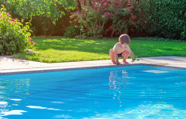 Little happy boy sitting on side of swimming pool in garden playing with his toy having fun. Summertime. Summer holidays.