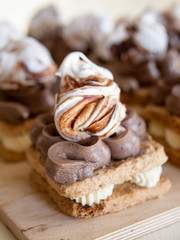 Portion cakes of meringue and cocoa butter cream in square form on a wooden stand, a piece of cake on a light wooden background. Restaurant serving. Selective focus, close up