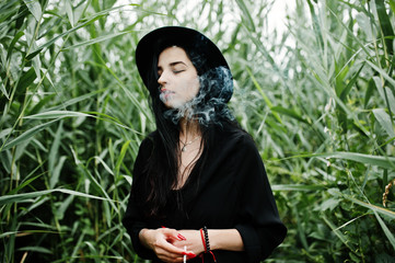 Sensual smoker girl all in black, red lips and hat. Goth dramatic woman smoking on common reed.