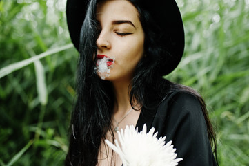 Sensual smoker girl all in black, red lips and hat. Goth dramatic woman hold white chrysanthemum...