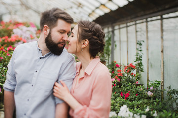 Young loving cheerful caucasian couple in glass greenhouse among colorful azalea flowers. Happy man and woman hugging and holding hands, spending time togehter.