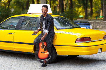 Handsome man with guitar in casual clothes near yellow taxi, sitting near door, posing confident....