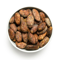 Roasted unpeeled cocoa beans in a white ceramic bowl isolated on white from above.