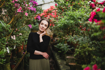 Young beautiful caucasian woman in glass greenhouse among colorful azalea flowers. Art portrait of a girl wearing classic fashionable clothes.