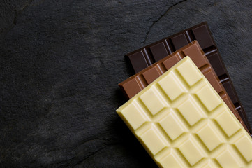 Slabs of white, milk and dark chocolate on top of each other on a black slate from above. Space for...