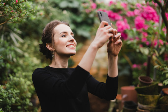 Young beautiful caucasian woman in glass greenhouse among colorful azalea flowers. Art portrait of a girl taking photo on her phone camera.