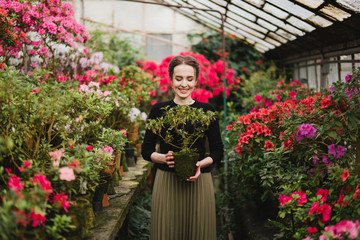 Young beautiful caucasian woman in glass greenhouse among colorful azalea flowers. Art portrait of a girl wearing classic fashionable clothes.