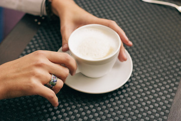 Woman with beautiful hands is drinking coffee in cafe. Close-up cappuccino in white cup, image with film effect and author processing.