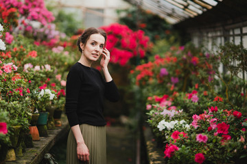 Young beautiful caucasian woman in glass greenhouse among colorful azalea flowers. Art portrait of a girl wearing classic fashionable clothes.