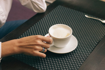 Woman with beautiful hands is drinking coffee in cafe. Close-up cappuccino in white cup, image with film effect and author processing.