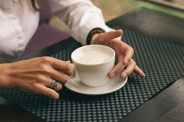 Woman with beautiful hands is drinking coffee in cafe. Close-up cappuccino in white cup, image with film effect and author processing.