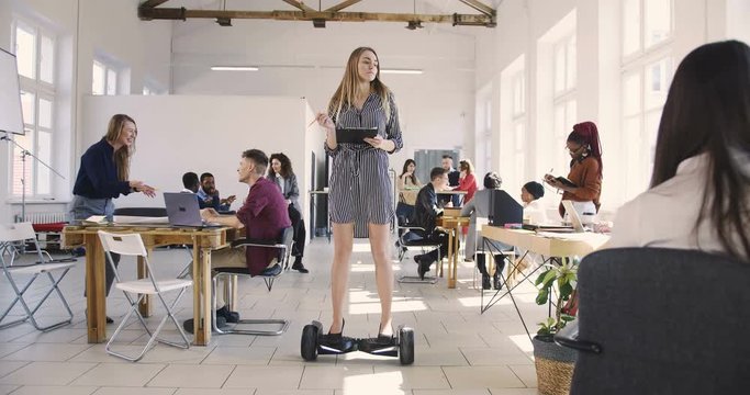 Young confident serious blonde business woman in dress riding electric gyroscooter in modern loft multiethnic office.