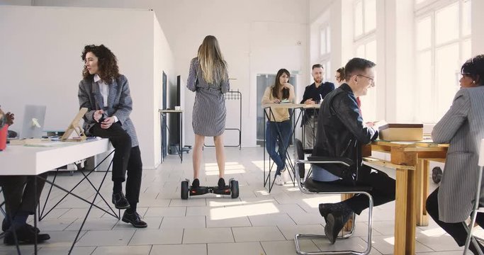 Camera folllows confident female leader using electric scooter to move around modern loft office. Healthy workplace.