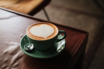 Cappuccino coffee with heart in turquoise cup and plate on wooden table. Warm toned filter effect. Coffee shop, barista latte art concept. Copyspace