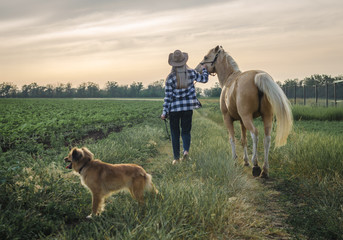 young blonde girl in a hat and a plaid shirt walks with a horse and dog on a farm in the village