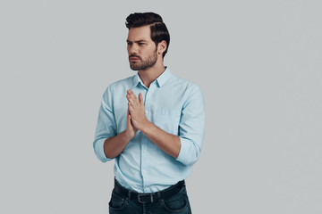 Taking time to make decision. Handsome young man looking away and keeping hands clasped while standing against grey background