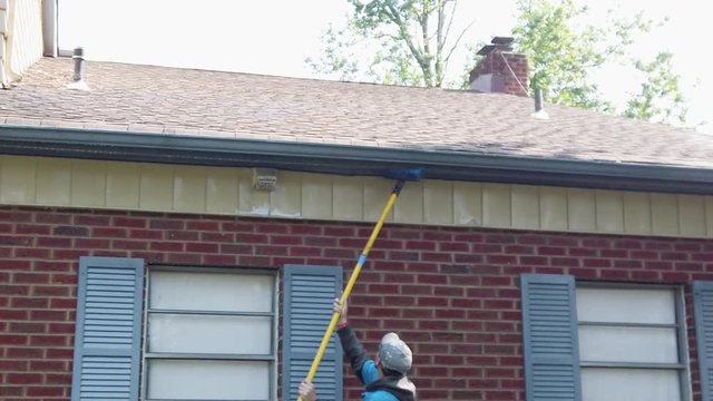 Male worker is cleaning the blue exterior soffit siding of a house with a scrub brush attached to a long pole. 