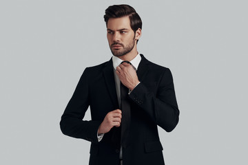 Everything must be perfect. Handsome young man in full suit adjusting tie and looking away while standing against grey background