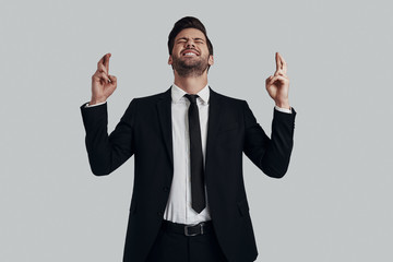 I wish? Handsome young man in full suit keeping fingers crossed and eyes closed while standing against grey background
