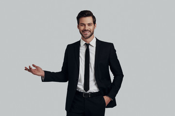 Just take a look! Handsome young man in full suit pointing copy space while standing against grey background