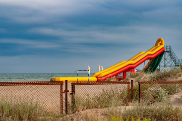 Obraz na płótnie Canvas Colorful inflatable slide on beach sand of coastline. Seascape with rusty metal fence and aquapark equipment on blue sky background. Landscape with nobody and picturesque cloudscape. Summer vacation 