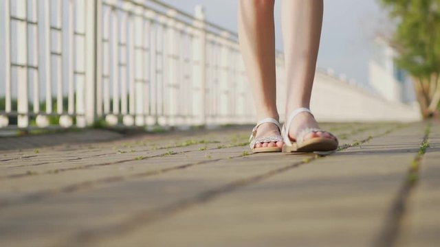 Woman walking on the bridge on the waterfront near the river in the summer, close-up of feet in sandals, slow motion, white sandals