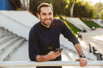 Handsome happy young strong sports man posing outdoors at the nature park location using mobile...