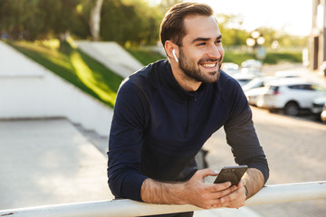 Happy young strong sports man posing outdoors at the nature park location using mobile phone...