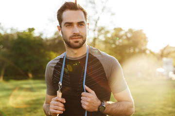 Handsome young strong sports man posing outdoors at the nature park location with skipping rope.