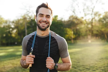 Gardinen Handsome young strong sports man posing outdoors at the nature park location with skipping rope. © Drobot Dean