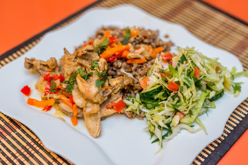 Pieces of rabbit stew with vegetables pepper, onion and herbs with boiled buckwheat and salad of fresh cucumbers and cabbage on a white plate. Mouth-watering delicious food at the gala dinner