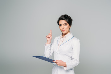 pretty mixed race doctor showing idea sign while holding clipboard isolated on grey